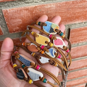 Op de afbeelding: Een set van zeven leren armbanden met kleurrijke keramische bedels. Elke armband heeft een andere bedel met een uniek patroon en kleurenschema. De bedels zijn bevestigd aan de leren banden met kleine metalen kralen.