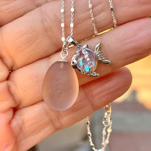 Puede incluir: Un collar de plata con dos colgantes: una piedra rosa translúcida en forma de lágrima y un dije en forma de pez con un centro iridiscente. La fina cadena de plata es visible. Joya.