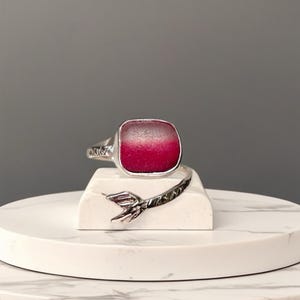 Puede incluir: Un anillo de plata con una piedra preciosa roja rectangular. El anillo presenta una banda decorativa y un diseño en forma de cola. El anillo se exhibe sobre una superficie de mármol blanco.