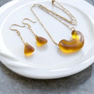 K&ouml;nnte beinhalten: Ein Goldcollier und Ohrringe mit Bernstein-Glasanh&auml;ngern. Die Halskette hat einen gro&szlig;en, gebogenen Bernstein-Glasanh&auml;nger, w&auml;hrend die Ohrringe kleinere, dreieckige Bernstein-Glasanh&auml;nger haben.