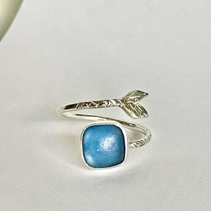 Puede incluir: Un anillo plateado con una piedra cuadrada de color azul claro. El anillo presenta un diseño en forma de hoja en un extremo y una banda texturizada. La piedra está engastada en un bisel cuadrado, creando un aspecto moderno y elegante.
