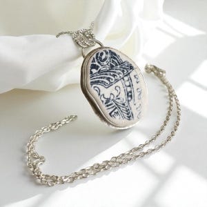 Puede incluir: Collar con colgante ovalado y cadena de plata. El colgante presenta una piedra color crema con un diseño en blanco y negro. El colgante está engastado en plata. El collar se muestra sobre un fondo blanco.