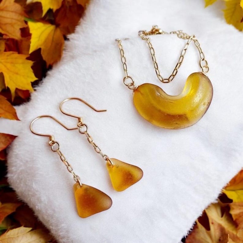 K&ouml;nnte beinhalten: Ein Set aus Bernstein-Schmuck mit einer Halskette und Ohrringen. Die Halskette hat einen gro&szlig;en, gebogenen Bernstein-Anh&auml;nger und eine goldene Kette. Die Ohrringe sind H&auml;ngeohrringe mit kleineren Bernstein-Tropfenanh&auml;ngern und goldenen Ketten.