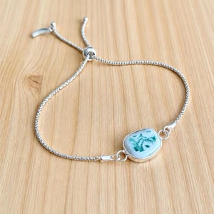 Puede incluir: Una pulsera de cadena tipo caja en tono plateado con un cierre deslizante ajustable y una piedra rectangular central. La piedra es blanca con inclusiones verdes similares al musgo, engastada en un marco de metal pulido en tono plateado.