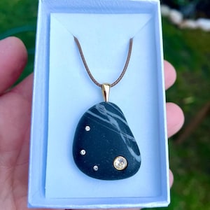 May include: A dark gray stone pendant with white veining, set in a gold-colored bail and strung on a brown cord. The pendant is adorned with three small clear crystals and one larger crystal. The pendant is presented in a white gift box.