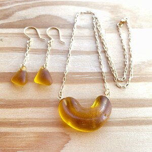 K&ouml;nnte beinhalten: Eine goldene Kettenkette mit einem braunen Glasanh&auml;nger in Form eines Halbmondes. Passende braune Glas-Dreiecksohrringe h&auml;ngen an goldenen Ketten.