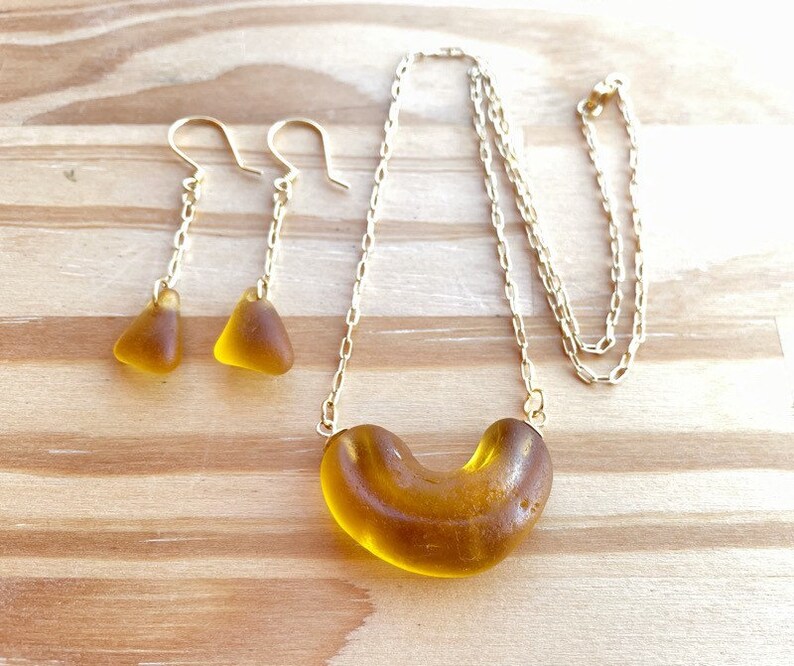 K&ouml;nnte beinhalten: Eine goldene Kettenkette mit einem braunen Glasanh&auml;nger in Form eines Halbmondes. Passende braune Glas-Dreiecksohrringe h&auml;ngen an goldenen Ketten.