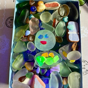 Puede incluir: Una caja azul llena de una variedad de coloridas piezas de vidrio de mar. El vidrio de mar es en tonos de verde, azul, transparente y marrón. Algunas piezas tienen forma de caras. La caja está sobre una superficie estampada.