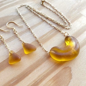K&ouml;nnte beinhalten: Eine goldene Kettenkette mit einem gro&szlig;en, bernsteinfarbenen Glasanh&auml;nger in Form eines Halbmondes. Passende Ohrringe mit kleineren, dreieckigen Bernstein-Glasst&uuml;cken h&auml;ngen an goldenen Ketten.