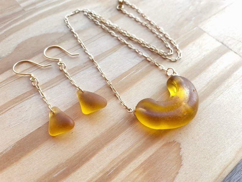 K&ouml;nnte beinhalten: Eine goldene Kettenkette mit einem gro&szlig;en, bernsteinfarbenen Glasanh&auml;nger in Form eines Halbmondes. Passende Ohrringe mit kleineren, dreieckigen Bernstein-Glasst&uuml;cken h&auml;ngen an goldenen Ketten.