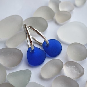 Puede incluir: Un par de pendientes de aro de plata con un encanto de vidrio de mar azul cobalto en cada aro.