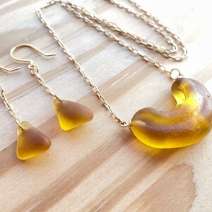 K&ouml;nnte beinhalten: Eine goldene Kettenkette mit einem gro&szlig;en, bernsteinfarbenen Glasanh&auml;nger in Form eines Halbmondes. Passende Ohrringe mit kleineren, dreieckigen Bernstein-Glasst&uuml;cken h&auml;ngen an goldenen Ketten.