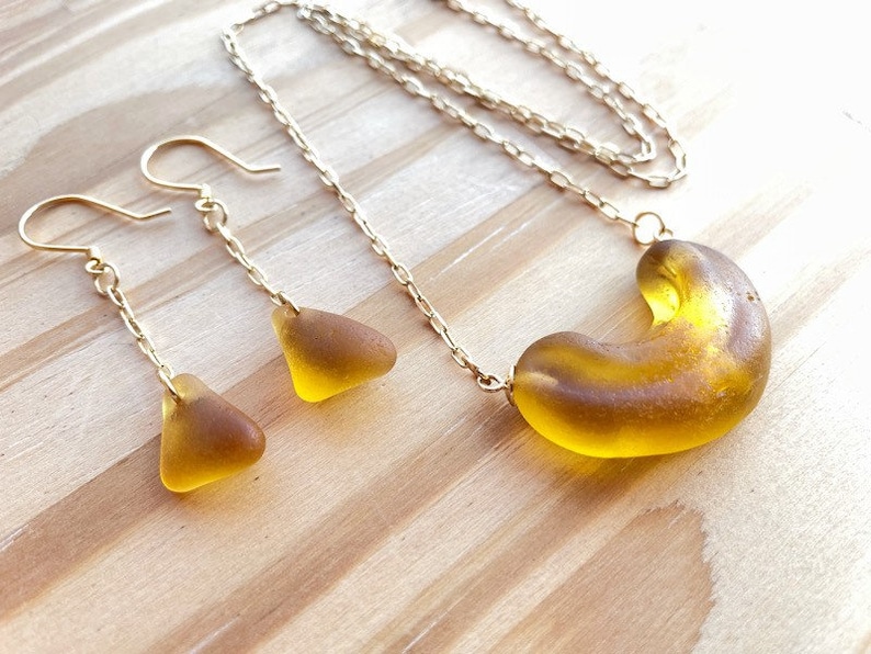 K&ouml;nnte beinhalten: Eine goldene Kettenkette mit einem gro&szlig;en, bernsteinfarbenen Glasanh&auml;nger in Form eines Halbmondes. Passende Ohrringe mit kleineren, dreieckigen Bernstein-Glasst&uuml;cken h&auml;ngen an goldenen Ketten.