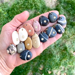 May include: A collection of small, smooth, oval-shaped river stone pendants in various colors including white, gray, black, tan, and pink. Each stone features a single, small, round, clear gemstone set in a gold-toned metal bezel and a gold jump ring.