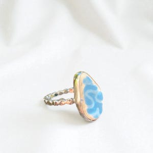 Puede incluir: Un anillo plateado con una piedra en forma de lágrima azul claro. La piedra tiene un borde blanco y un diseño azul en espiral. La banda del anillo tiene un diseño texturizado con cuentas.