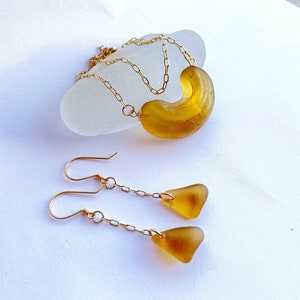 K&ouml;nnte beinhalten: Eine goldene Kettenkette mit einem gro&szlig;en Anh&auml;nger aus bernsteinfarbenem Meerglas. Passende Ohrringe mit kleineren Anh&auml;ngern aus bernsteinfarbenem Meerglas h&auml;ngen an goldenen Ketten.