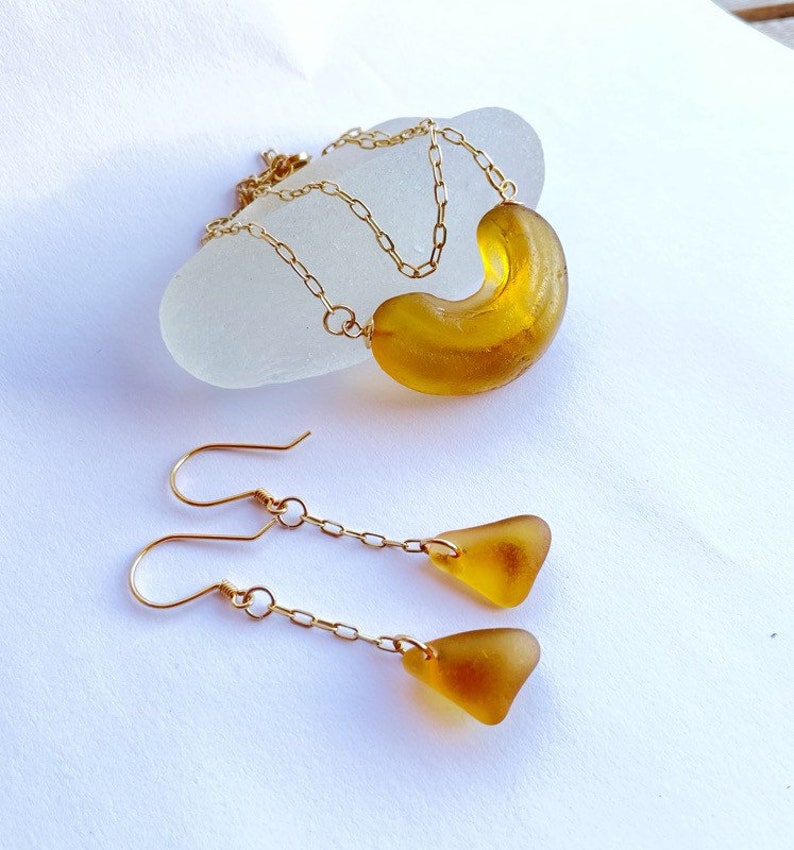 K&ouml;nnte beinhalten: Eine goldene Kettenkette mit einem gro&szlig;en Anh&auml;nger aus bernsteinfarbenem Meerglas. Passende Ohrringe mit kleineren Anh&auml;ngern aus bernsteinfarbenem Meerglas h&auml;ngen an goldenen Ketten.
