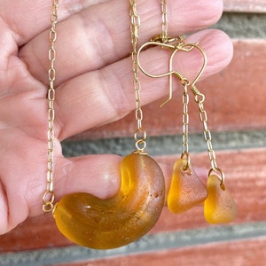 K&ouml;nnte beinhalten: Ein Set aus Bernstein-Meerglasschmuck mit einer Halskette und Ohrringen. Die Halskette hat ein gro&szlig;es, gebogenes St&uuml;ck Meerglas und die Ohrringe haben zwei kleinere St&uuml;cke Meerglas. Der Schmuck ist an goldenen Ketten aufgef&auml;delt.