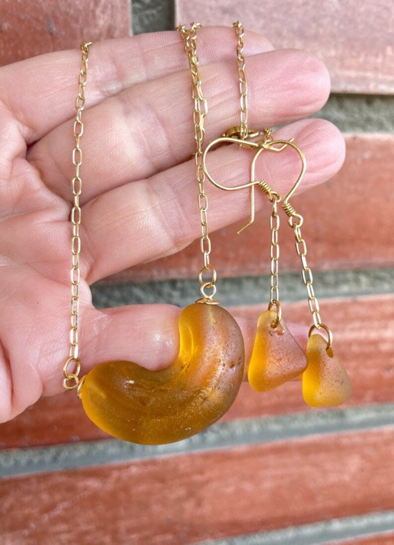 K&ouml;nnte beinhalten: Ein Set aus Bernstein-Meerglasschmuck mit einer Halskette und Ohrringen. Die Halskette hat ein gro&szlig;es, gebogenes St&uuml;ck Meerglas und die Ohrringe haben zwei kleinere St&uuml;cke Meerglas. Der Schmuck ist an goldenen Ketten aufgef&auml;delt.