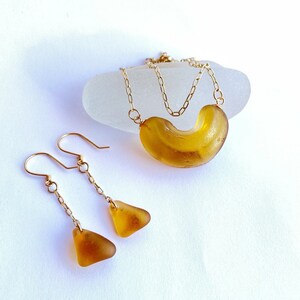 K&ouml;nnte beinhalten: Ein Set aus Bernstein-Meerglasschmuck mit einer Halskette und Ohrringen. Die Halskette hat eine goldene Kette und ein gro&szlig;es, gebogenes St&uuml;ck Meerglas. Die Ohrringe sind H&auml;ngeohrringe mit goldenen Ketten und kleineren dreieckigen Meerglasscheiben.