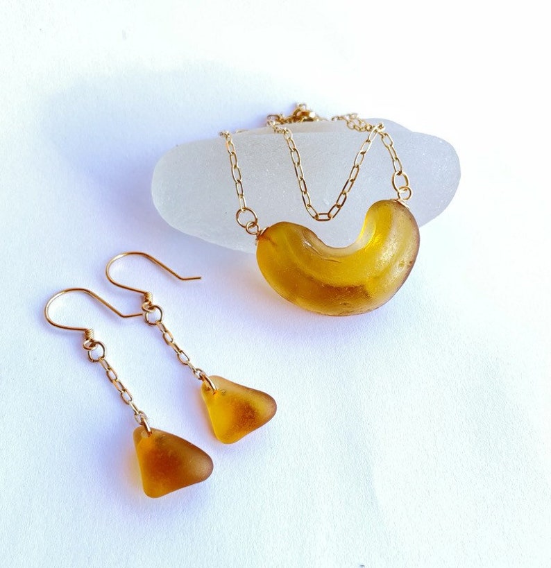 K&ouml;nnte beinhalten: Ein Set aus Bernstein-Meerglasschmuck mit einer Halskette und Ohrringen. Die Halskette hat eine goldene Kette und ein gro&szlig;es, gebogenes St&uuml;ck Meerglas. Die Ohrringe sind H&auml;ngeohrringe mit goldenen Ketten und kleineren dreieckigen Meerglasscheiben.