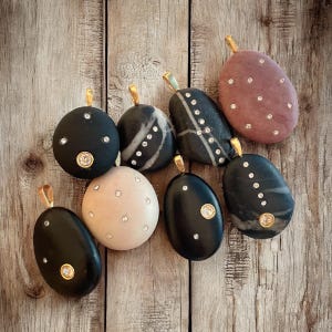 May include: A collection of eight smooth, rounded stone pendants, each featuring a gold-toned metal bail. The stones vary in color, including black, beige, and mauve, and are embellished with small, clear, sparkling inset crystals.