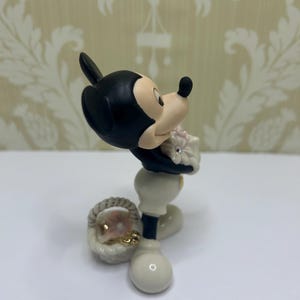 May include: A figurine of Mickey Mouse in a classic pose, holding a bouquet of pink flowers. The figure is primarily black, white, and beige, with a small basket at his feet. The figurine has a glossy finish.
