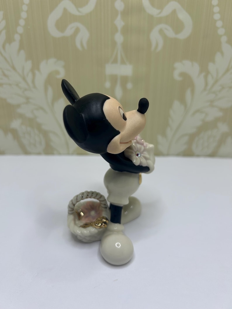 May include: A figurine of Mickey Mouse in a classic pose, holding a bouquet of pink flowers. The figure is primarily black, white, and beige, with a small basket at his feet. The figurine has a glossy finish.
