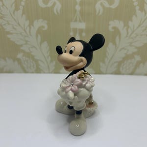 May include: A figurine of Mickey Mouse, featuring black ears, a tan face, and white gloves and shoes. Mickey is holding a bouquet of pink flowers with a gold ring and a small diamond. The figurine is set against a patterned background.