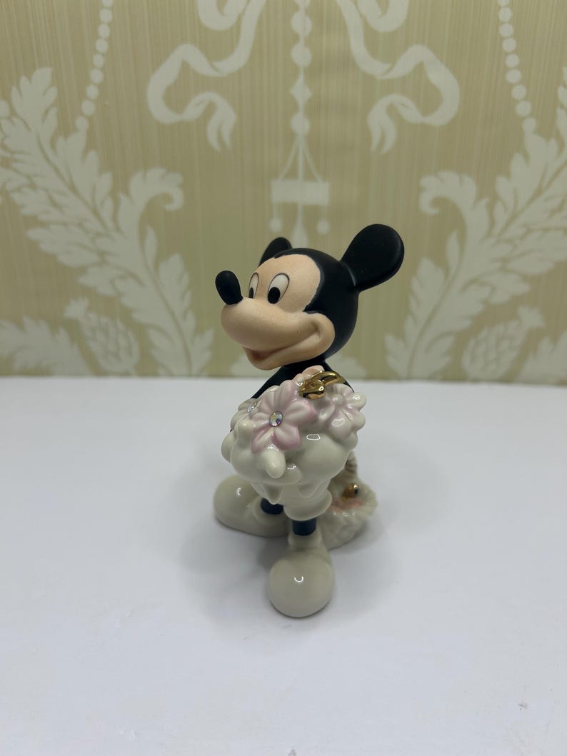 May include: A figurine of Mickey Mouse, featuring black ears, a tan face, and white gloves and shoes. Mickey is holding a bouquet of pink flowers with a gold ring and a small diamond. The figurine is set against a patterned background.
