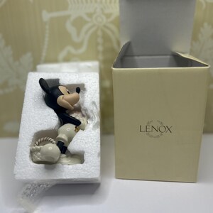 May include: A black, white, and tan Mickey Mouse figurine in a protective foam insert, next to a beige Lenox box with the lid open. The figurine is holding a white basket. The box has the Lenox logo.