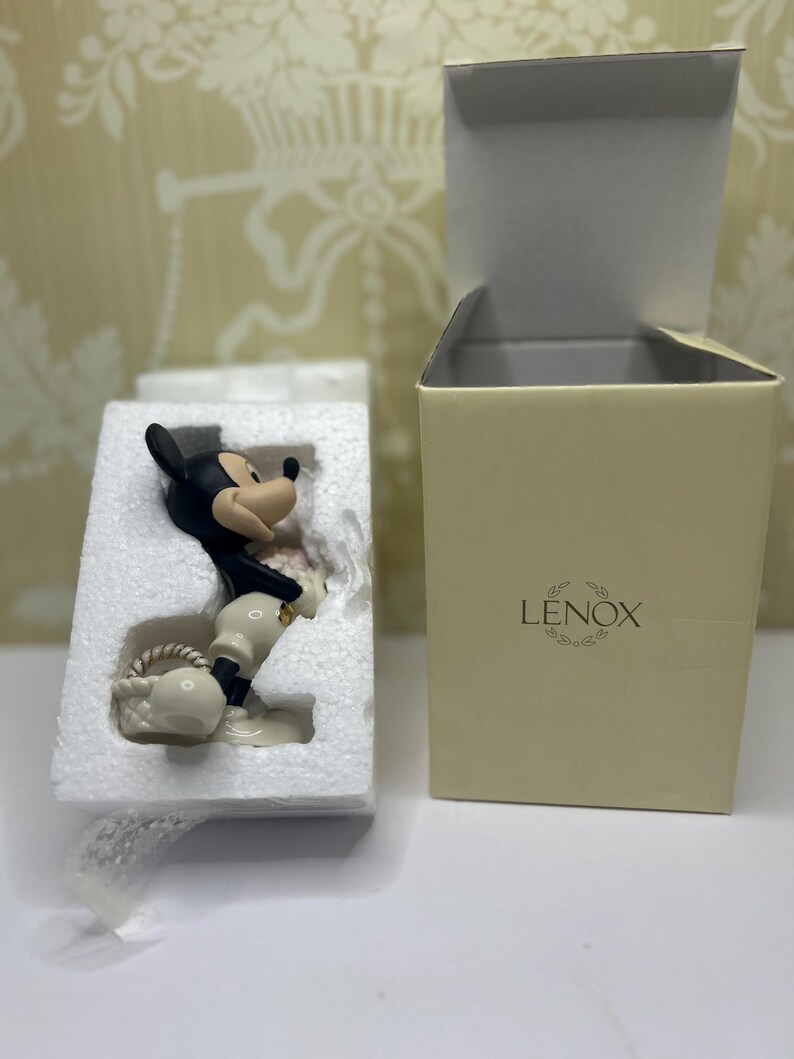 May include: A black, white, and tan Mickey Mouse figurine in a protective foam insert, next to a beige Lenox box with the lid open. The figurine is holding a white basket. The box has the Lenox logo.