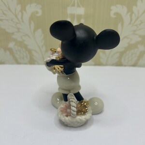 May include: A black and white Mickey Mouse figurine holding a basket with pink and gold accents. The figurine has a black head with large ears, a white body, and black shorts. The basket is white with a woven handle.