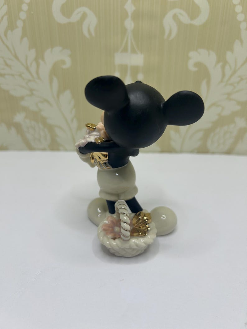 May include: A black and white Mickey Mouse figurine holding a basket with pink and gold accents. The figurine has a black head with large ears, a white body, and black shorts. The basket is white with a woven handle.