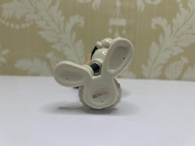 May include: A ceramic figurine of a cartoon character, viewed from below. The figure has large, rounded ears, a black nose, and a white body. The base is marked with "LENOX DISNEY".