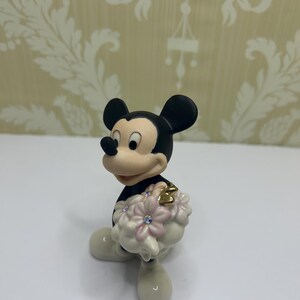 May include: A figurine of Mickey Mouse in a black outfit with white gloves and shoes. Mickey is holding a bouquet of pink flowers with crystal accents and a gold heart. The figurine is on a white surface.