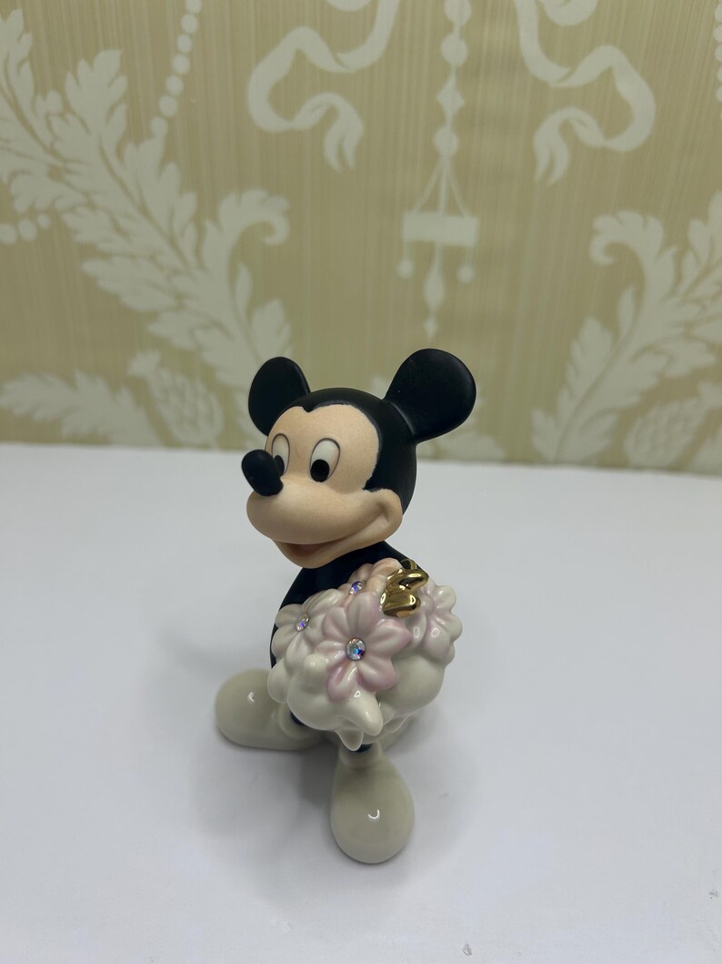 May include: A figurine of Mickey Mouse in a black outfit with white gloves and shoes. Mickey is holding a bouquet of pink flowers with crystal accents and a gold heart. The figurine is on a white surface.