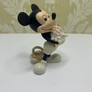 May include: A figurine of Mickey Mouse holding a bouquet of pink flowers with rhinestone accents and a small basket. Mickey is wearing white gloves, pants, and shoes, with a black body and ears. The figurine has a glossy finish.