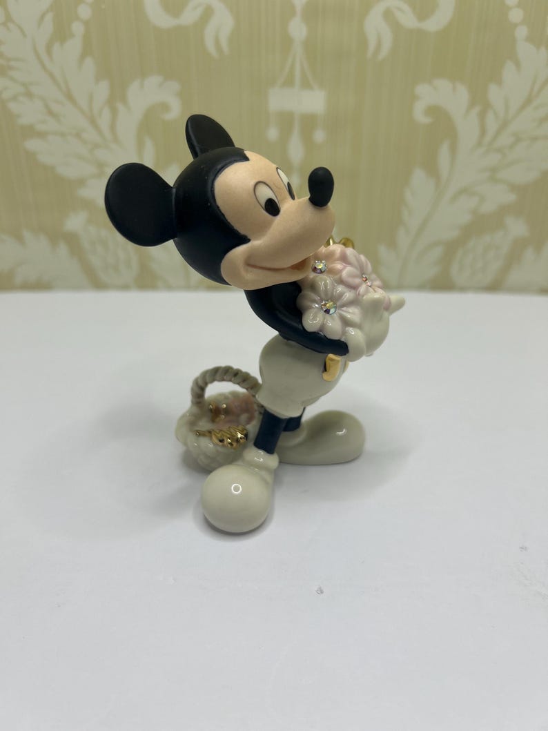 May include: A figurine of Mickey Mouse holding a bouquet of pink flowers with rhinestone accents and a small basket. Mickey is wearing white gloves, pants, and shoes, with a black body and ears. The figurine has a glossy finish.