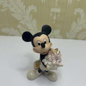 May include: A figurine of Mickey Mouse holding a bouquet of pink flowers with blue and clear crystal accents. Mickey is wearing white gloves, shoes, and a black outfit. The figurine has a glossy finish.