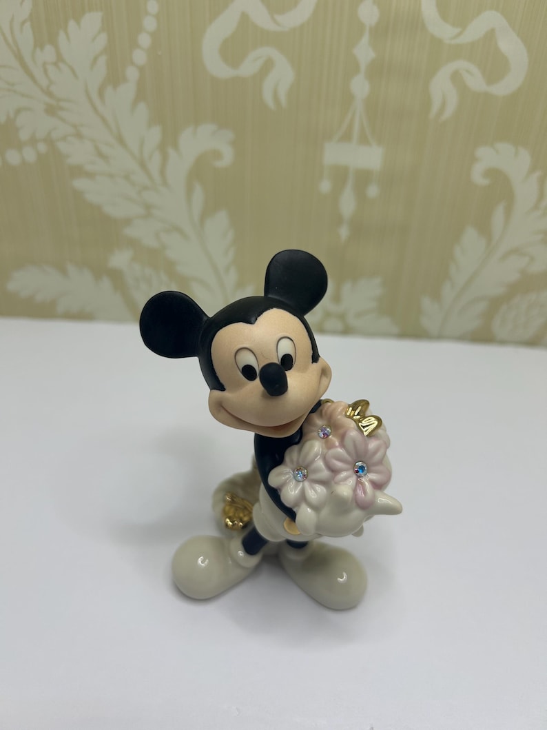 May include: A figurine of Mickey Mouse holding a bouquet of pink flowers with blue and clear crystal accents. Mickey is wearing white gloves, shoes, and a black outfit. The figurine has a glossy finish.