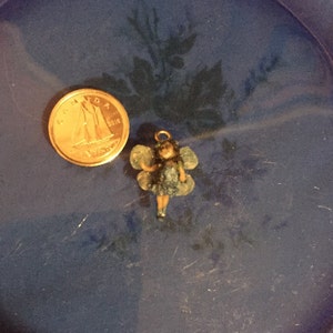May include: A small blue fairy charm with wings and a gold-colored head. The charm is sitting on a blue surface.