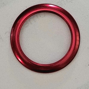 May include: A close-up of a shiny, red ring. The ring is circular with a smooth, reflective surface. It appears to be made of metal. The background is a light, textured surface. The ring is the focal point of the image.