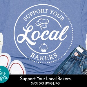 May include: A blue t-shirt with a white circle design that says "Support Your Local Bakers" with a chef's hat, cupcake, and the word "Local" in a stylized font.