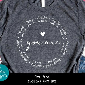 May include: A gray t-shirt with a white circle design that says "you are" with a heart in the center. The circle is surrounded by words and bible verses including "Strong", "Amazing", "Capable", "Chosen", "Never Alone", "Enough", "Created", "Beautiful", "Victorious", "Always Loved".