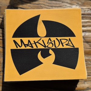May include: Square sticker with a yellow background and a black graphic design. The design includes a stylized symbol and the word "MAKISUPA" in black lettering. The sticker is on a wooden surface.