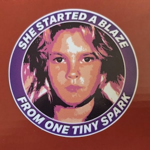 May include: A round sticker with a portrait of a person in shades of pink, orange, and white, surrounded by a purple ring. The text reads "SHE STARTED A BLAZE FROM ONE TINY SPARK."