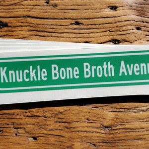 May include: A stack of white rectangular stickers with a green and white border. The text "Knuckle Bone Broth Avenue" is printed in white on the green background. The stickers are on a wooden surface.