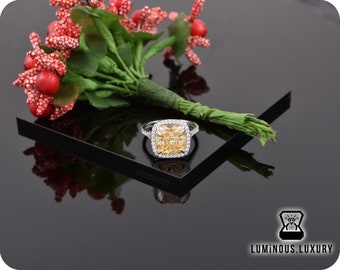 May include: A square-shaped, yellow gemstone ring with a halo of smaller diamonds set in a silver band. The ring is on a black surface with a bouquet of red berries and green leaves in the background.