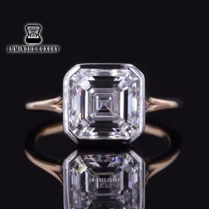 May include: A square-shaped diamond engagement ring with a rose gold band. The diamond is set in a bezel setting with a square shape. The ring is on a black background.